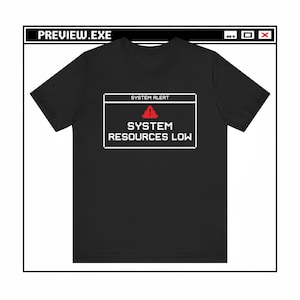 May include: Black t-shirt with a white pixelated graphic resembling a computer error message. The message reads "SYSTEM ALERT" above a red triangle, followed by "SYSTEM RESOURCES LOW". The top of the graphic reads "PREVIEW.EXE".