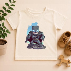 May include: Cream-colored t-shirt featuring a cartoon illustration of Batman in a dark gray armored suit. The graphic includes a blue background and is centered on the shirt. Brown leather shoes and a wooden horse toy are next to the shirt.