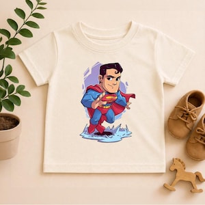 May include: Off-white short-sleeved t-shirt featuring a cartoon illustration of Superman. The superhero is depicted in his classic blue and red costume, with a red cape, and the Superman logo on his chest. The shirt is made of a soft material.