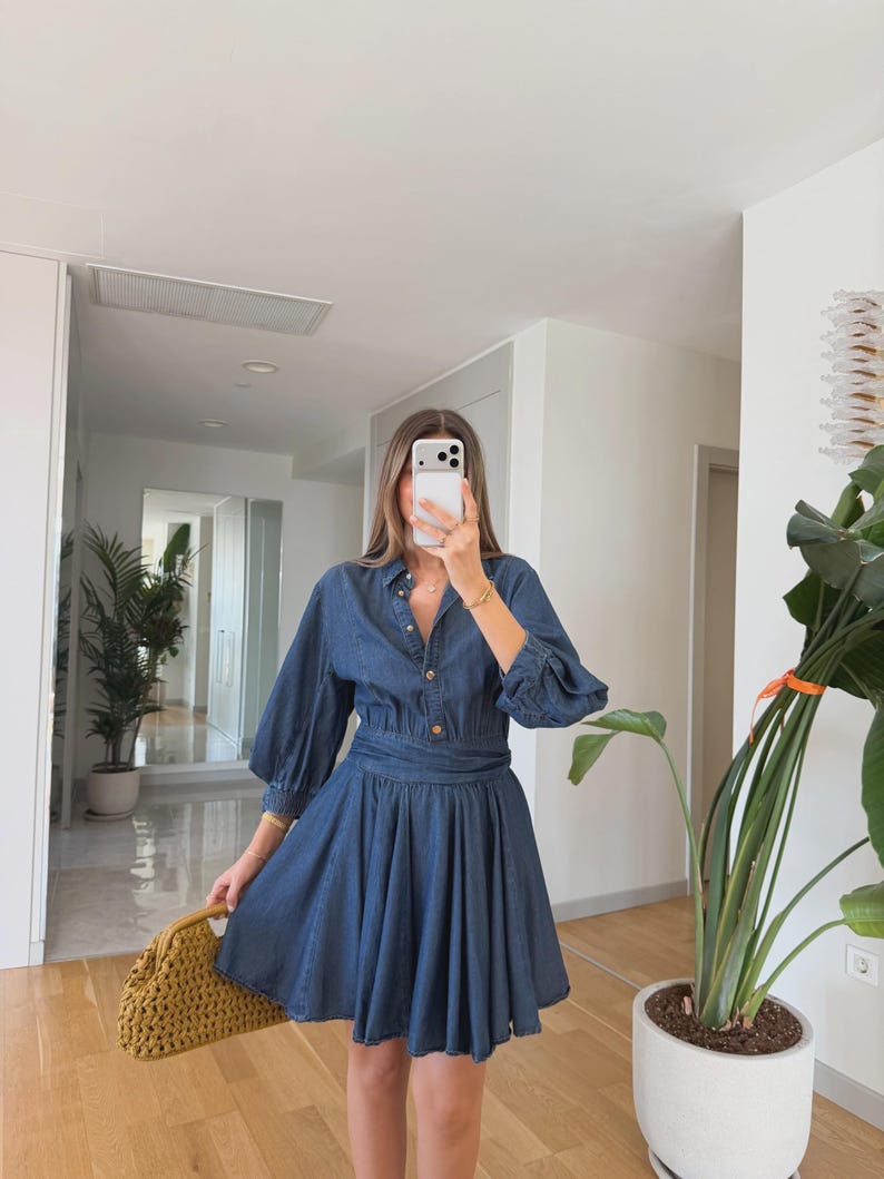May include: A blue denim dress featuring a button-down top and a flared skirt. The dress has long sleeves and a cinched waist. A yellow woven handbag is held in the hand. The dress is a medium blue colour.