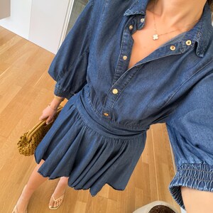 May include: A blue denim dress with a button-down front, short sleeves, and a flared skirt. The dress has gold-coloured buttons and a cinched waist. The person is wearing gold jewellery and sandals, and carrying a woven handbag.