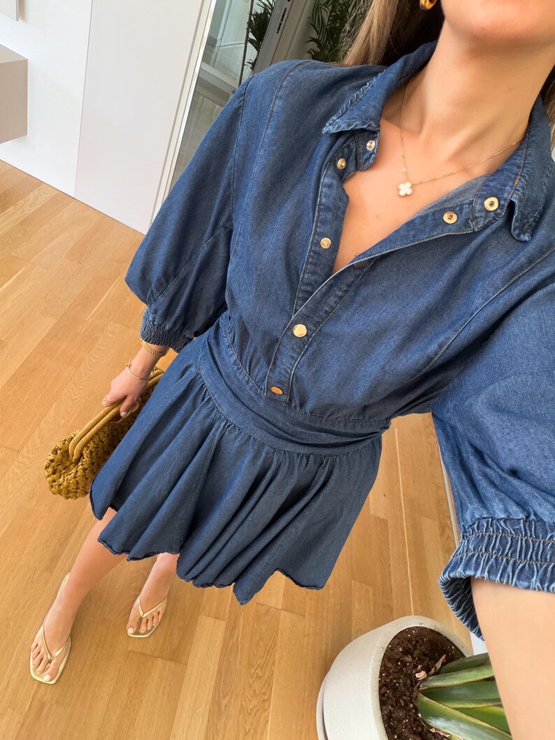 May include: A blue denim dress with a button-down front, short sleeves, and a flared skirt. The dress has gold-coloured buttons and a cinched waist. The person is wearing gold jewellery and sandals, and carrying a woven handbag.