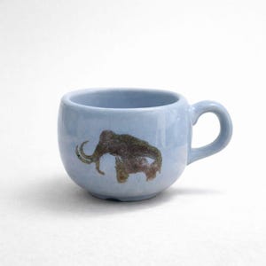Könnte beinhalten: Hellblaue Keramik-Tasse mit runder Form und Henkel. Auf der Vorderseite befindet sich eine braune und beige Mammut-Illustration. Geeignet für Kaffee, Tee oder andere Getränke. Ungefähr 8 cm hoch.