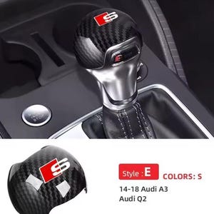 May include: Carbon fiber style gear shift knob with a red and white "S" logo. The knob is shown installed in a car's interior, with a silver metal shaft and black leather boot. A second knob is shown below.