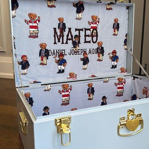 Pode incluir: Um baú de armazenamento azul-claro com fechos de metal dourado e um cadeado em forma de coração. O interior é forrado com tecido às riscas brancas e azuis com um padrão de ursinhos de peluche e o nome MATEO DANIEL JOSEPH AGOS.