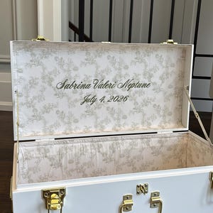 May include: A white rectangular storage trunk with gold-toned metal latches and a handle. The interior is lined with a light-coloured fabric featuring a delicate grey floral pattern. The text "Sabrina Valeri Neptune" and "July 4, 2026" is printed.