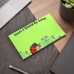 May include: A bright green notepad with the text "Stuff I Should Be Doing" in dark blue. The notepad features a cartoon ladybug and blue floral accents. A pen, laptop, and notebook are in the background.