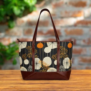 May include: A tote bag featuring a black background with a repeating pattern of cream and orange dandelion-like circular motifs. The bag has dark brown leather-like handles and a matching brown base, with a zipper closure along the top edge.