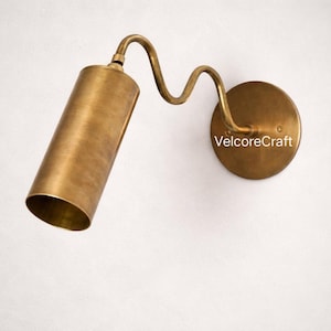 Puede incluir: Aplique de pared de latón con una pantalla cilíndrica y un brazo curvo. El aplique está montado sobre una base redonda y tiene el texto "VelcoreCraft". El acabado es un dorado metálico cálido.