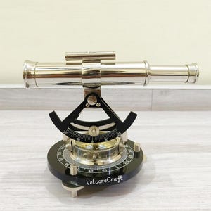 May include: A polished brass nautical telescope with a black base and compass. The telescope is mounted on a black base with a compass and a degree scale. The text "VelcoreCraft" is printed on the base.