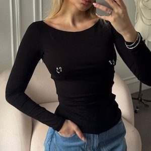 May include: A black long-sleeve top with a scoop neckline and two silver-colored decorative accents. The top is paired with blue jeans. The person in the image is holding a smartphone.
