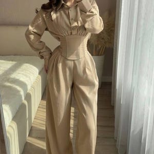 May include: A beige two-piece outfit consisting of a button-down shirt, a corset, and wide-leg pants. The shirt has long sleeves and a collared neckline. The corset cinches the waist, and the pants have a relaxed fit. The outfit is made of a light fabric.