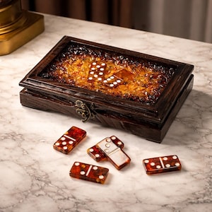 May include: A wooden domino set with a hinged lid and metal clasp. The interior is filled with amber-colored glass pieces. Several dominoes with white dots are scattered around the box on a marble surface. The dominoes are red-brown with white dots.