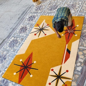 May include: A rectangular area rug featuring a mustard yellow background with cream-colored geometric shapes. The design includes black starburst motifs, orange diamond accents, and small red circular dots arranged in a pattern.