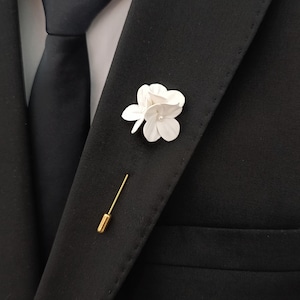 Op de afbeelding: Een witte bloemspeld met een parel in het midden is vastgemaakt aan een zwart colbert. Een goudkleurige speld is eronder geplaatst. Een zwarte stropdas en een wit overhemd zijn ook zichtbaar.