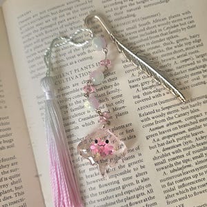 May include: A handmade bookmark with a silver feather design, adorned with pink and white beads and a pink tassel. A clear charm with a pink flower design is attached, resting on an open book.