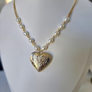 May include: A gold heart-shaped locket necklace with a floral pattern. The locket is suspended from a chain with small, white, oval pearls. The necklace is displayed on a white mannequin.