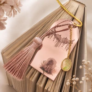 Könnte beinhalten: Ein bogenförmiges Lesezeichen in Roségold mit einer zartrosa Quaste und einem goldenen Metallhaken. Die Oberfläche zeigt zarte Illustrationen von Kürbissen und Kerzen. Eine kleine goldene Kette mit einem ovalen, irisierenden Steinanhänger ist befestigt.