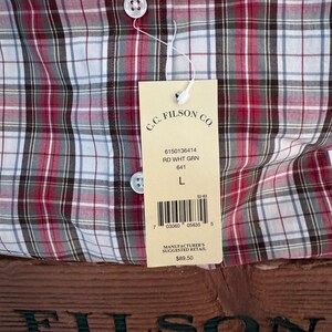 May include: A red, white, and green plaid button-up shirt with a tag from C.C. Filson Co. The tag includes the text "RD WHT GRN" and "L". The shirt is displayed on a wooden surface with the Filson logo.