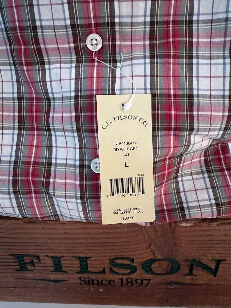 May include: A red, white, and green plaid button-up shirt with a tag from C.C. Filson Co. The tag includes the text "RD WHT GRN" and "L". The shirt is displayed on a wooden surface with the Filson logo.