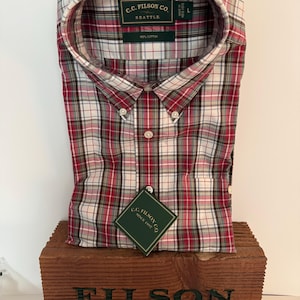 May include: A red, white, and black plaid button-down shirt with the text "C.C. Filson Co. Seattle" on the label. The shirt is made of 100% cotton and is displayed on a wooden box with the text "FILSON Since 1897".