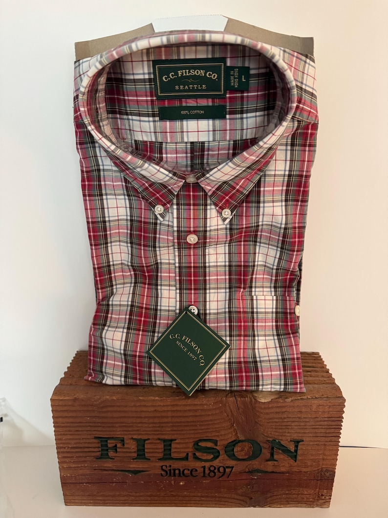 May include: A red, white, and black plaid button-down shirt with the text "C.C. Filson Co. Seattle" on the label. The shirt is made of 100% cotton and is displayed on a wooden box with the text "FILSON Since 1897".