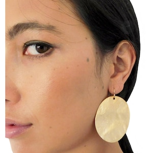 May include: Close-up of a person's face with a large, round, gold-coloured earring. The earring is a flat disc shape with a textured surface. The earring hangs from a small gold hook. The person has dark hair and fair skin.