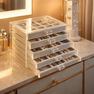 Może przedstawiać: Przezroczysty akrylowy organizer na biżuterię z wieloma szufladami i przegródkami, wystawiony na białej komodzie. Organizer jest wypełniony różnymi elementami biżuterii. Widoczny jest również biały marmurowy stojak na biżuterię i butelki perfum.