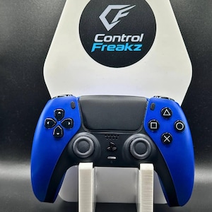 May include: A blue and black games controller on a white stand. The controller has a black centre and blue sides with black buttons. The stand has the text "Control Freakz" on it.