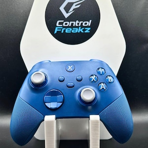 May include: A blue games controller with white thumbsticks and buttons, on a white stand. The controller has textured grips and the letters X, A, and B are visible. The background features a white stand with the Control Freakz logo.