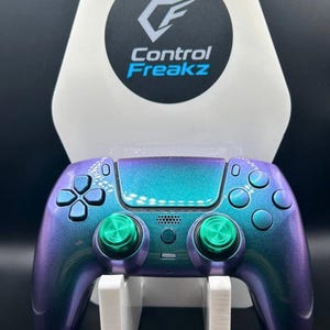 May include: A custom video game controller with a colour-shifting purple and teal finish. The controller has green thumbsticks and buttons. It is displayed on a white stand with the "Control Freakz" logo.