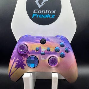 May include: An Xbox controller with a sunset-themed design, including palm trees and city outlines. The controller has a colour scheme of purple, pink, and gold, with blue buttons and thumbsticks. It is displayed on a white stand, with the Control Freakz logo.
