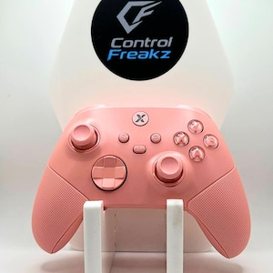 May include: A pink games controller with textured grips, resting on a white stand. The controller has buttons and joysticks. Above it is a white and black logo that says "Control Freakz".