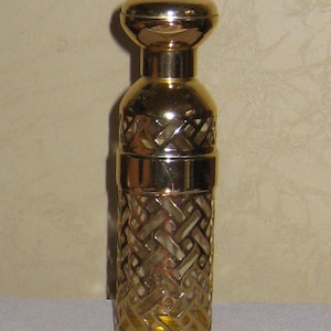 Puede incluir: Un frasco de perfume dorado con un patrón decorativo tejido en la parte inferior. El frasco tiene una parte superior redondeada y un cuerpo cilíndrico. El frasco es de metal y tiene un acabado brillante.