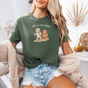 Op de afbeelding: Groen t-shirt met de tekst "Off to be brave" en een illustratie van twee stripfiguren. Het shirt wordt gecombineerd met een spijkerbroek. Een beige vest wordt over de schouders gedragen.