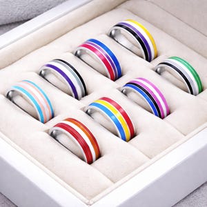 Könnte beinhalten: Eine Sammlung von Edelstahlringen mit bunten Emaille-Streifen, präsentiert in einer weißen Schmuckschatulle. Die Ringe weisen verschiedene Farbkombinationen auf, darunter Regenbogen, Rosa, Blau, Schwarz und Weiß. Die Ringe sind ein Modeaccessoire.