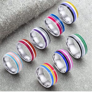 May include: A selection of stainless steel rings with vibrant enamel stripes. Each ring showcases a unique colour combination, such as red, blue, yellow, pink, and green. The rings have a polished, reflective surface.