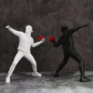 Puede incluir: Esculturas de dos figuras lanzando flores rojas. Una figura está vestida de blanco, con una sudadera con capucha y una máscara. La otra figura está vestida de negro, con un bombín y ropa oscura. El fondo es gris oscuro.