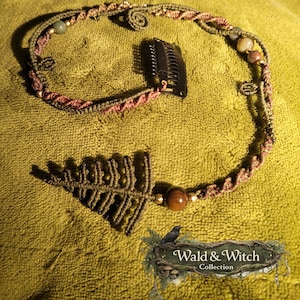 Könnte beinhalten: Ein handgefertigtes Makramee-Haaraccessoire mit Blattdesign und schwarzem Clip. Das Haarteil hat grüne und rotbraune Kordeln, verziert mit Perlen. Das Logo der Wald & Witch Collection ist sichtbar.