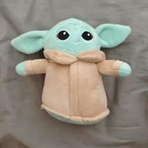May include: A soft toy of a character with large ears, big black eyes, and a light green complexion. The character is wearing a beige coat with a light green trim. The toy is on a grey surface.