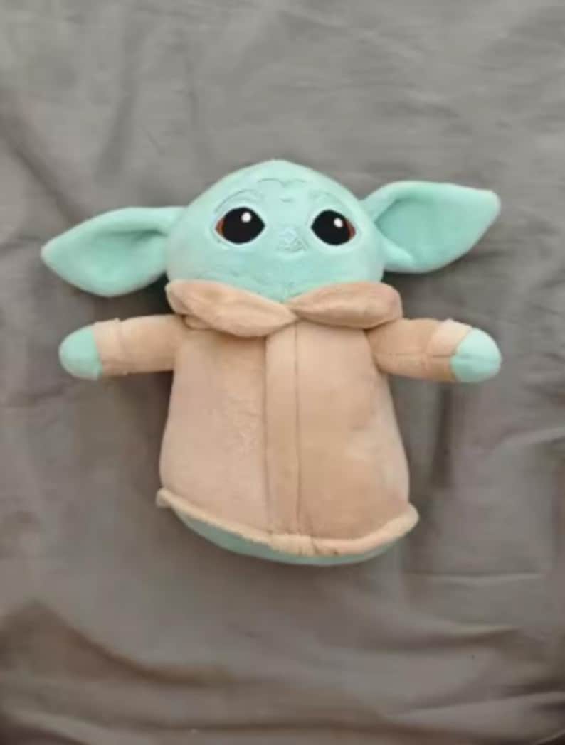 May include: A soft toy of a character with large ears, big black eyes, and a light green complexion. The character is wearing a beige coat with a light green trim. The toy is on a grey surface.