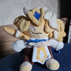 Puede incluir: Un peluche de un personaje con cabeza amarilla, pelo blanco y un atuendo azul y blanco. El juguete tiene una cara sonriente y est&aacute; sentado sobre una superficie azul. El personaje tiene un emblema azul en la cabeza.