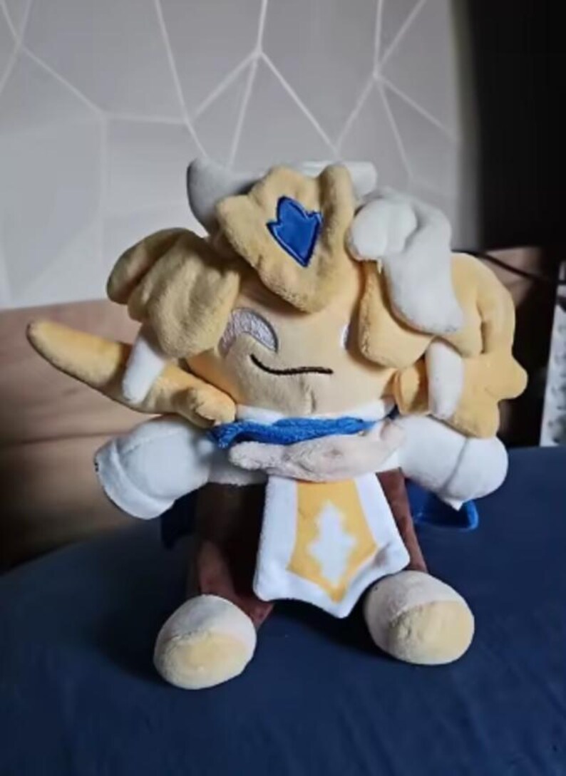 Puede incluir: Un peluche de un personaje con cabeza amarilla, pelo blanco y un atuendo azul y blanco. El juguete tiene una cara sonriente y est&aacute; sentado sobre una superficie azul. El personaje tiene un emblema azul en la cabeza.