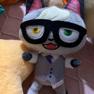 Może przedstawiać: Pluszowa zabawka przedstawiająca kota z szarym futrem, okularami w czarnych oprawkach i szarą kamizelką na białej koszuli i fioletowym krawacie. Kot ma jedno żółte i jedno zielone oko. To postać z Animal Crossing.