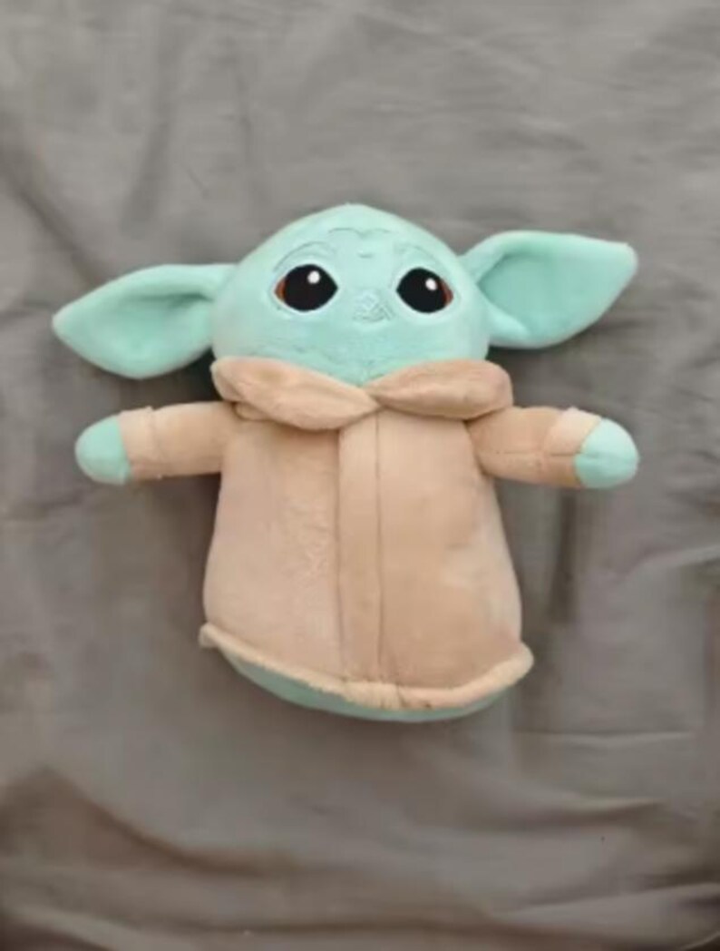 May include: A soft toy of a character with large ears, big black eyes, and a light green complexion. The character is wearing a beige coat with a light green trim. The toy is on a grey surface.