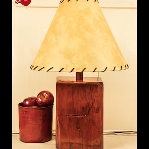 May include: A table lamp with a rectangular, dark reddish-brown wooden base and a conical, cream-colored lampshade featuring dark brown stitched edges. Beside the lamp sits a cylindrical, deep red container holding several shiny red apples.