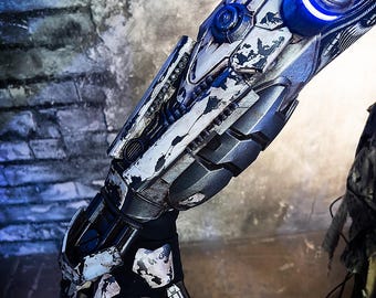 May include: A detailed view of a robotic arm with a distressed, metallic finish. The arm features a combination of silver, black, and white paint, with blue illuminated accents. The design includes intricate mechanical details and a futuristic aesthetic.