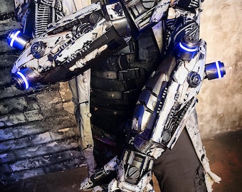 May include: A detailed view of a futuristic robotic arm and hand, painted in white and black with blue glowing accents. The design features intricate mechanical details and a distressed finish, suggesting a worn or battle-damaged aesthetic. The costume is part of a larger sci-fi ensemble.