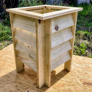 May include: A wooden planter box with a square top and slatted sides. The planter is made from light-coloured wood and has four legs. The design incorporates a slatted pattern on the sides, allowing for ventilation. Suitable for outdoor use, it is measured in centimetres.
