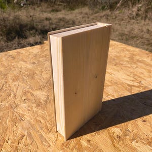 Op de afbeelding: Een lichtgekleurde, houten doos in de vorm van een boek. De doos is gemaakt van onbewerkt hout, met zichtbare nerf en een eenvoudig, rechthoekig ontwerp. De doos staat rechtop op een spaanplaat oppervlak.